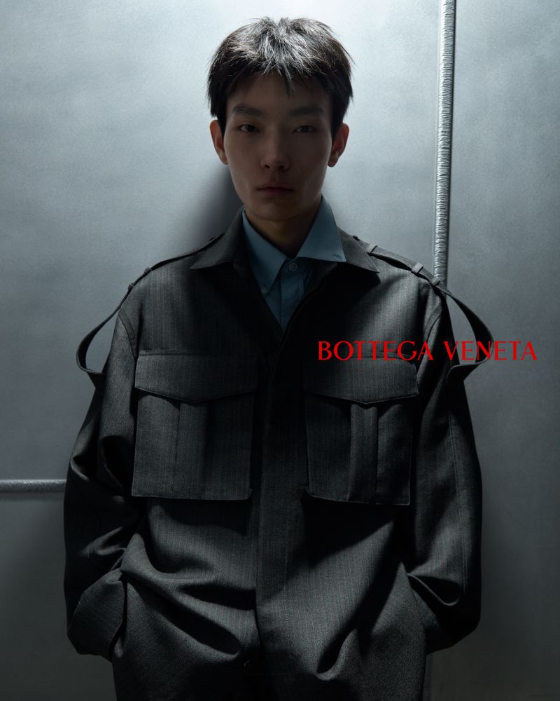 East Asian male model posing in a charcoal utility jacket and pale blue collared shirt against a cool grey studio backdrop.