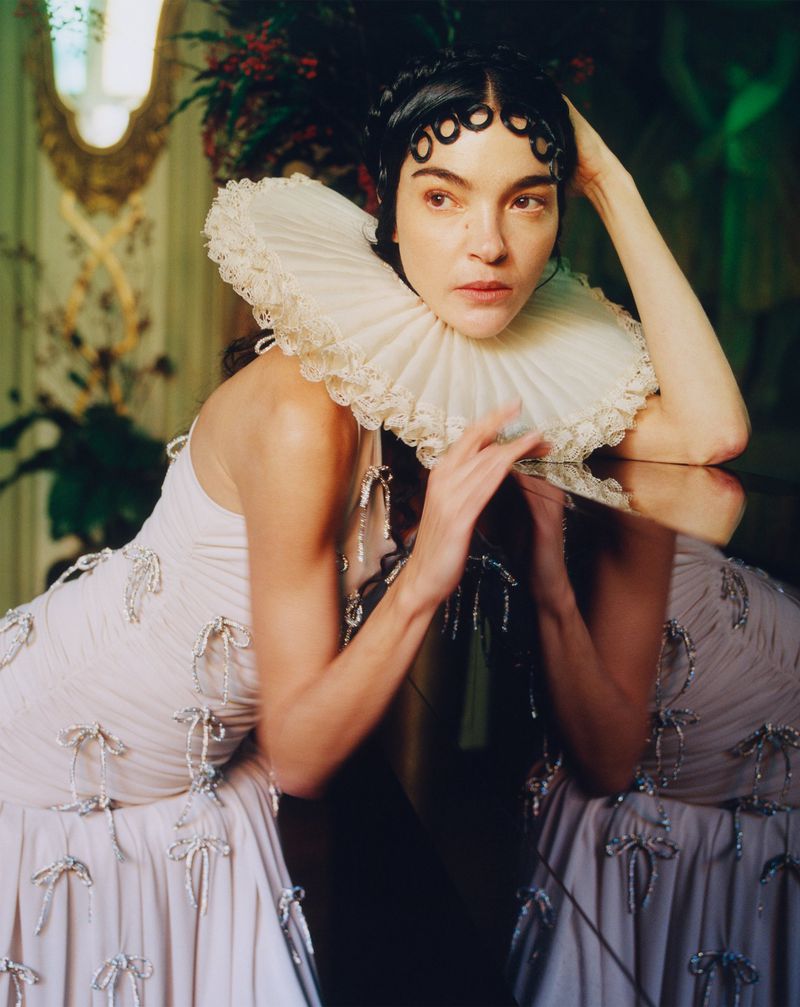 Adult female model with dark hair poses, wearing a ruffled white collar and sleeveless dress, against an ornate indoor backdrop.