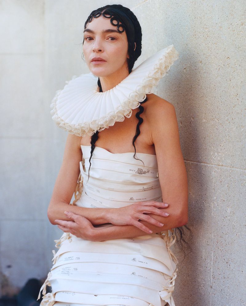 Woman in a paper dress with a large white ruffled collar stands against a textured wall in a fashion forward editorial style portrait.