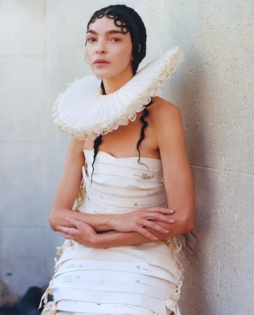 Woman in a paper dress with a large white ruffled collar stands against a textured wall in a fashion forward editorial style portrait.