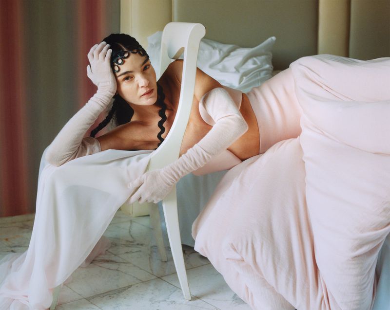 Woman in a light pink dress sitting on a white chair in a bedroom setting with light pink bedding, wearing gloves, and a braided hairstyle.