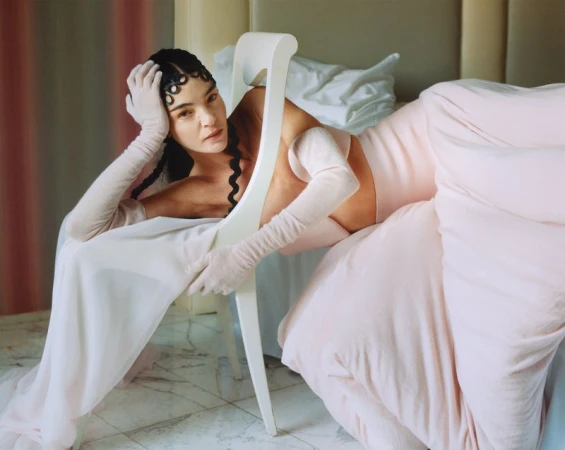 Woman in a light pink dress sitting on a white chair in a bedroom setting with light pink bedding, wearing gloves, and a braided hairstyle.