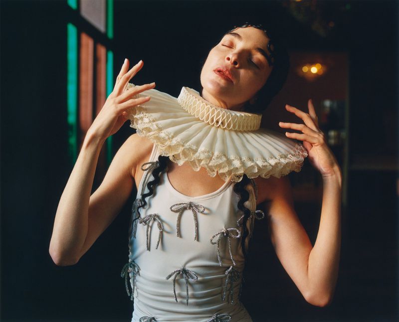 Woman in a white dress with silver bows and a ruffled collar poses indoors with hands raised toward her face, bathed in bright light.