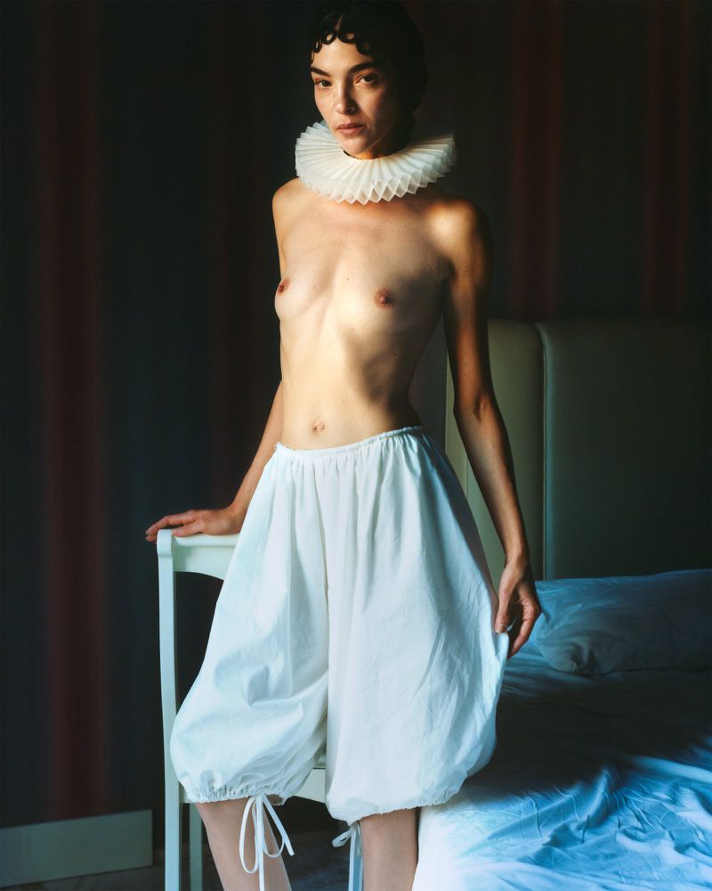 Artistic studio shot of a slender woman wearing a ruffled neckpiece and white bloomers, posing against a dark wall and bedroom setting.