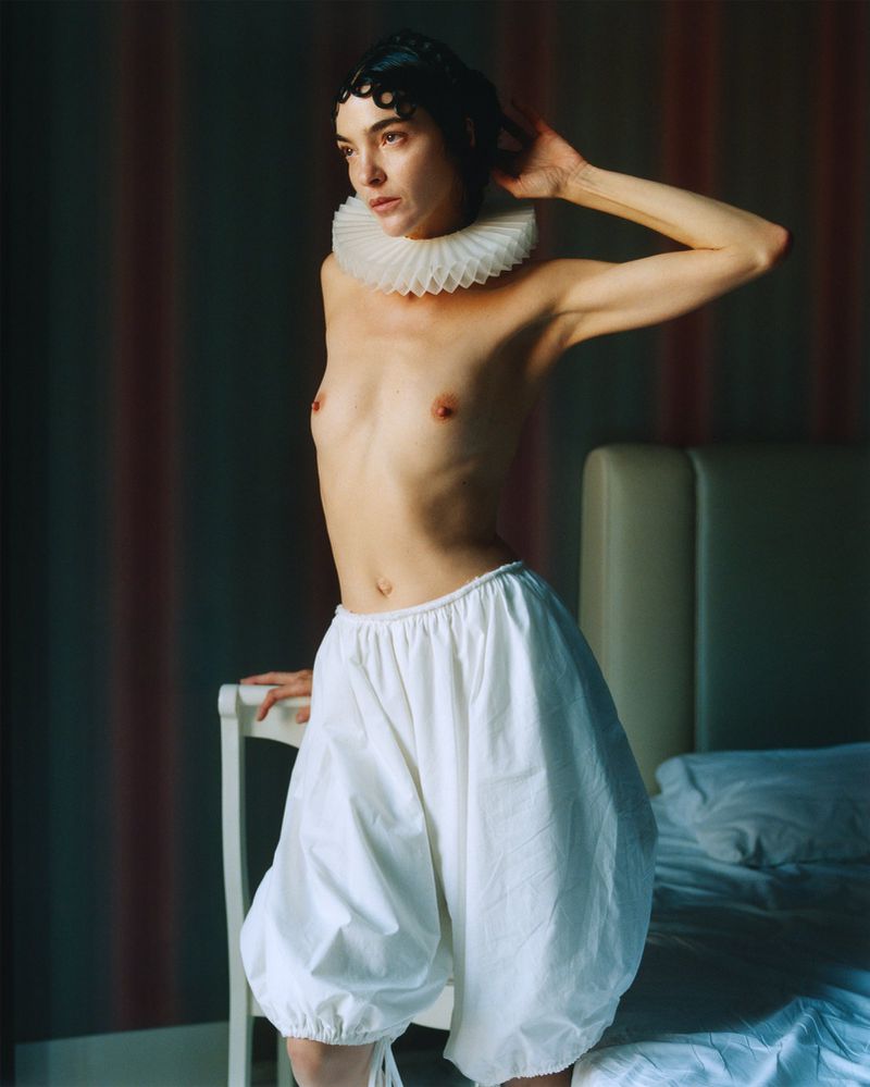 Adult woman in puffy white pants, wearing a white ruffled collar, resting hand on a white chair in an indoor bedroom setting.