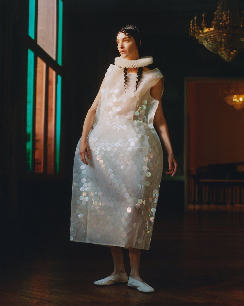 Adult female in a white dress with ruffled collar stands in an indoor setting with a chandelier and large window in the background.