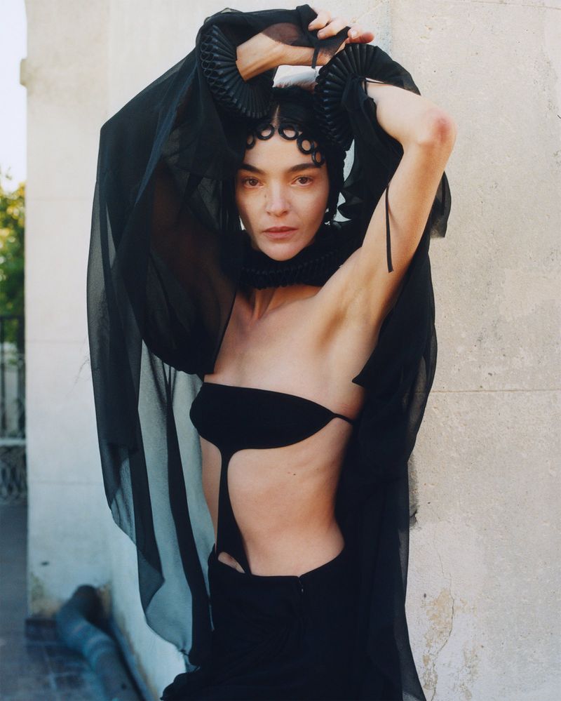 Adult female fashion model in black strapless dress, long black gloves, and sheer black veil with horn accents poses outdoors.