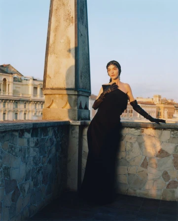 Elegant woman in black gown and long gloves on rooftop terrace with city view, bathed in warm, late-day sunlight.