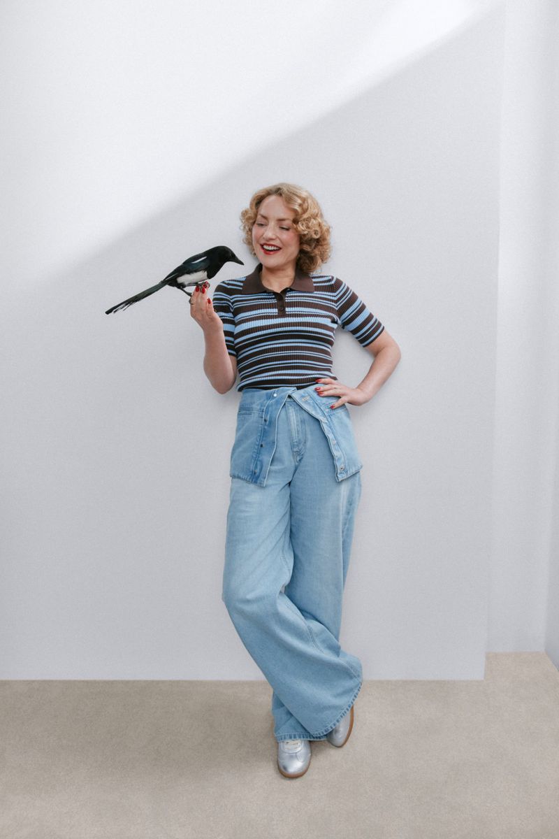 Woman with curly blonde hair wearing a striped knit polo and wide-leg jeans holding a perched magpie bird against a white wall.