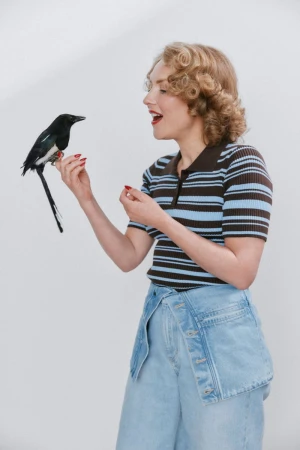 A woman with blonde curly hair wearing a blue and brown striped polo shirt and denim jeans holding a black and white magpie bird.
