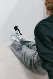 Person seated on the floor wearing grey denim and white sneakers with a magpie perched on their shoe.