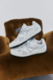 Pair of white and silver Axel Arigato sneakers stacked on a tufted brown velvet armchair against a plain white studio backdrop.