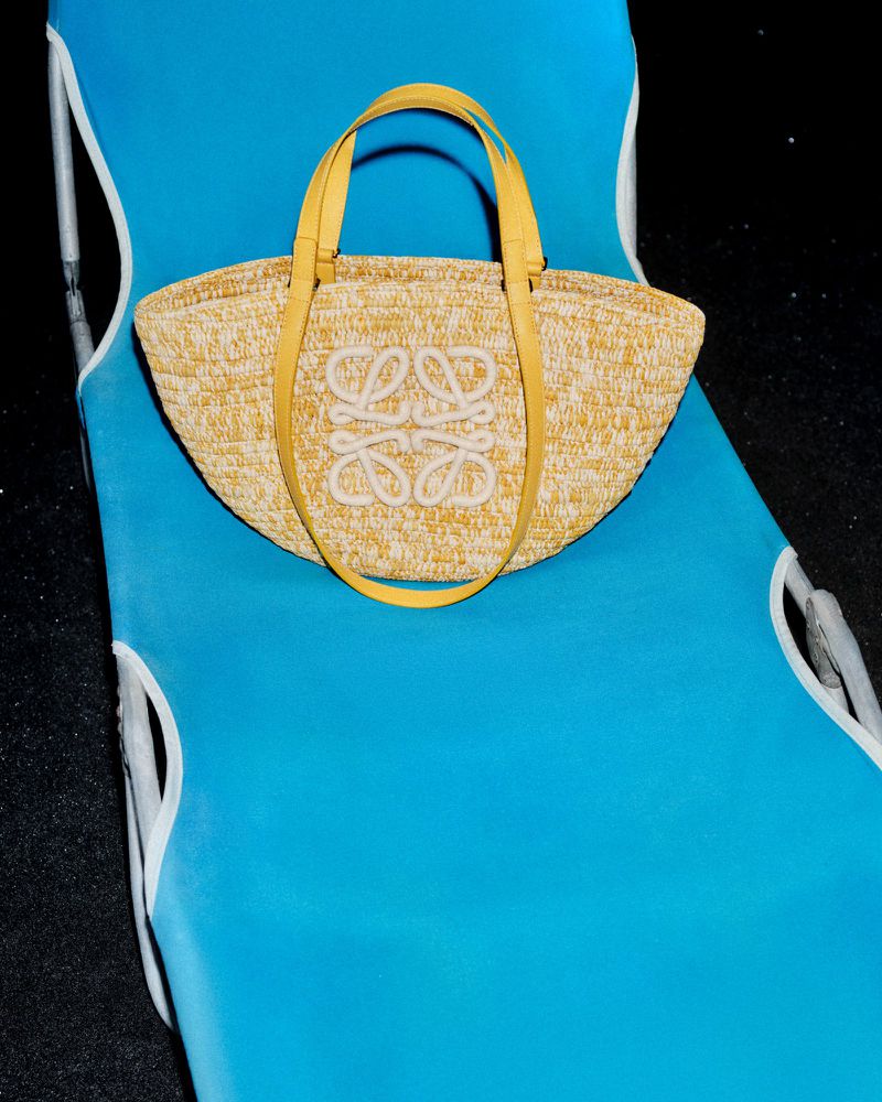 A Loewe straw basket bag with yellow leather handles and a centered anagram logo rests on a bright blue fabric pool lounge chair.