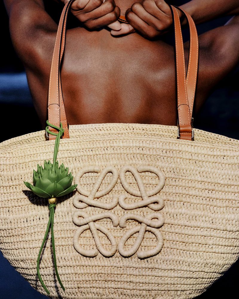 Woven raffia tote bag with leather straps and green charm held against a model's bare back.