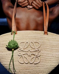 Woven raffia tote bag with leather straps and green charm held against a model's bare back.