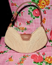 Loewe crescent-shaped woven raffia handbag with a brown leather patch and strap set against a pink floral printed textile.