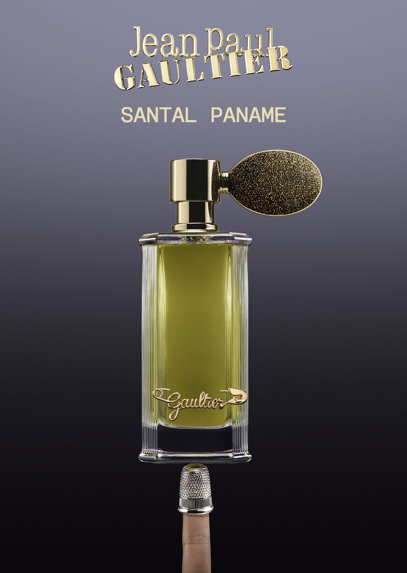 A rectangular Jean Paul Gaultier Santal Paname perfume bottle balanced on a silver thimble atop a finger against a dark gradient background.