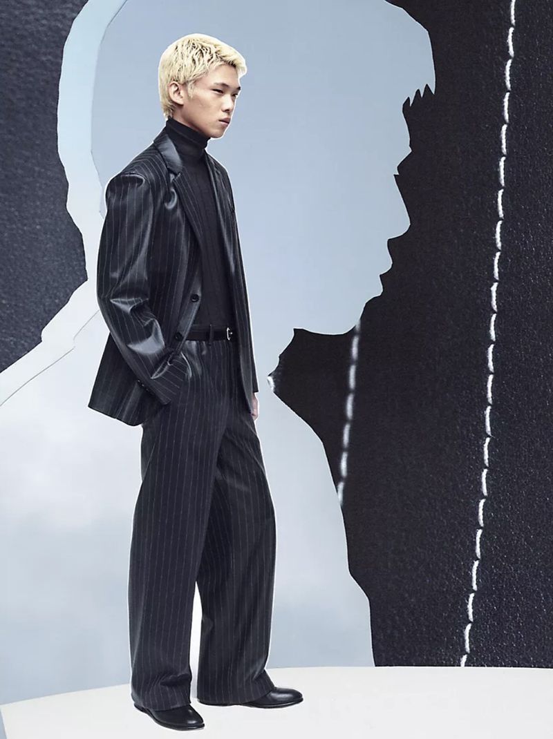 A man with bleached blonde hair wearing a pinstripe blazer and trousers stands against a stylized cut-out silhouette backdrop.