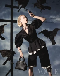 Model posing in a black puff-sleeve peplum jacket and fringe-trimmed shorts while black crows fly around a utility pole.