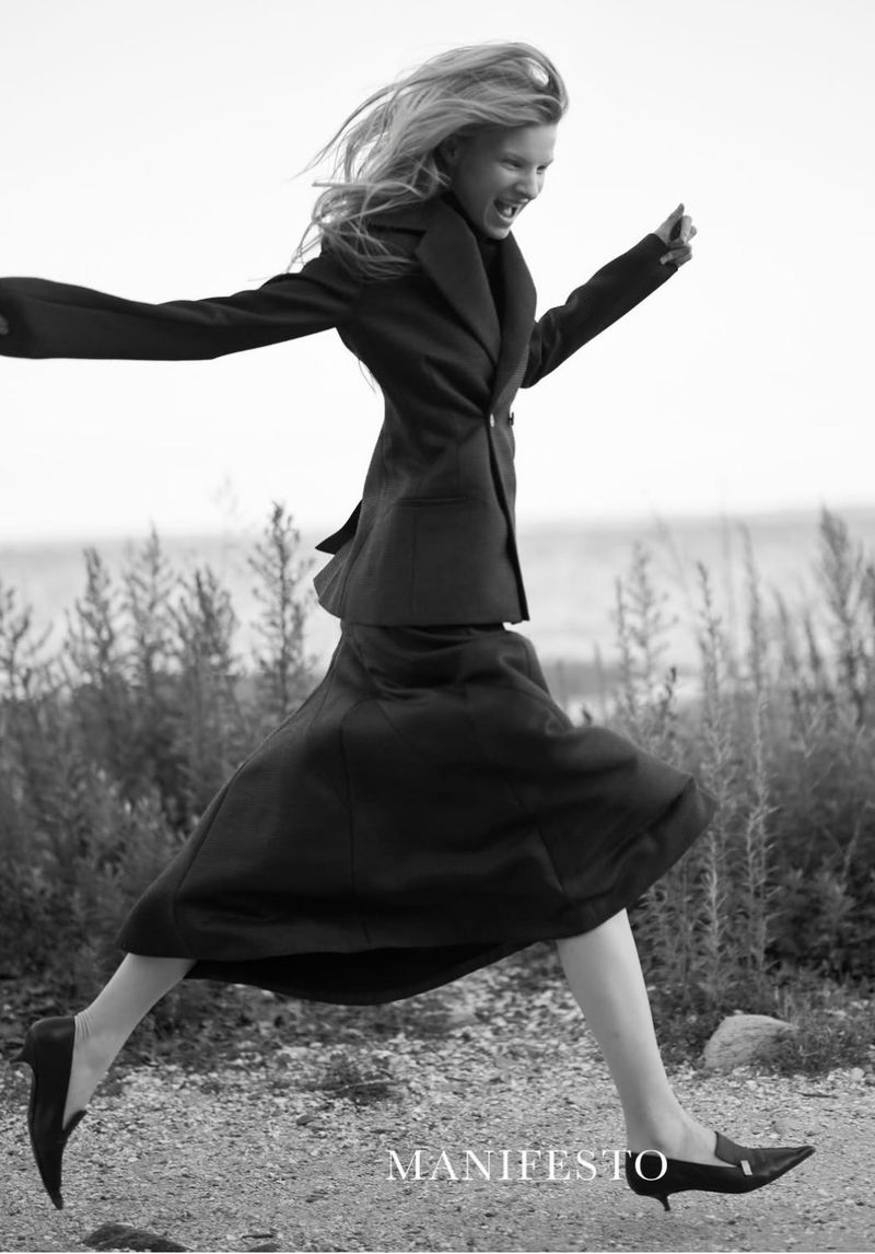 A blonde model wearing a dark tailored blazer and flared midi skirt leaps across a rocky, grassy landscape in a black and white editorial.