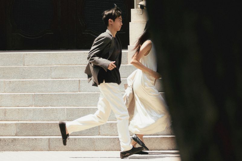 An East Asian man running down stone steps in a blazer and white trousers alongside Mika Schneider in a white dress.