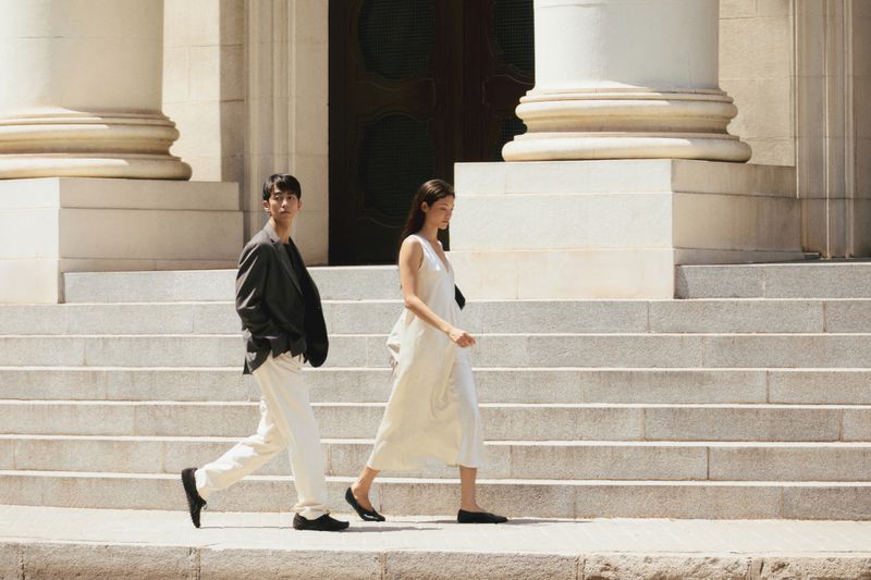 Model Mika Schneider in a white slip dress walking alongside a man in a dark blazer on stone steps.