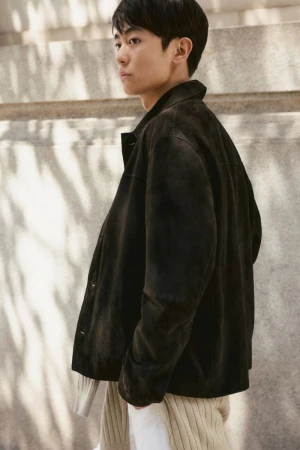 An East Asian man wearing a dark brown suede jacket standing in dappled sunlight against a neutral stone wall.