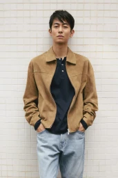 East Asian man posing in a camel suede jacket, navy polo shirt, and light blue jeans against a grid tiled wall.