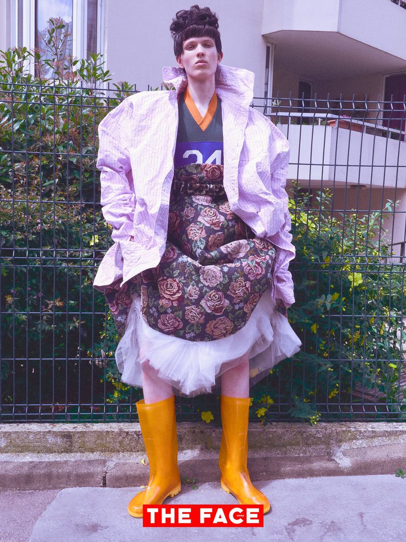 Model wearing an oversized lavender coat, layered floral skirt, and bright yellow rain boots standing before a fenced hedge.