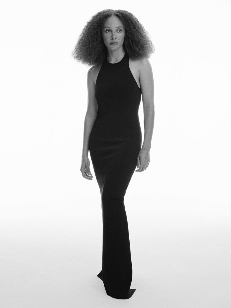 Elegant full-length portrait of an adult woman with curly hair wearing a black halter dress against a bright, neutral background in a studio.