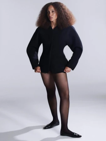 Woman with Afro wearing black jacket, tights, and flats poses in a full shot against a gradient gray background in a fashion studio.