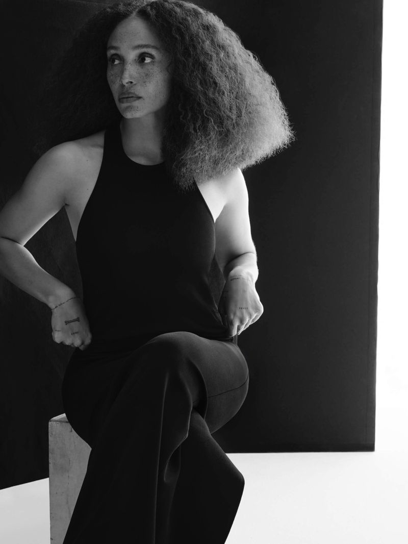 Portrait of a Black female in a black tank top and slacks, sitting on a concrete block against a black and white background.