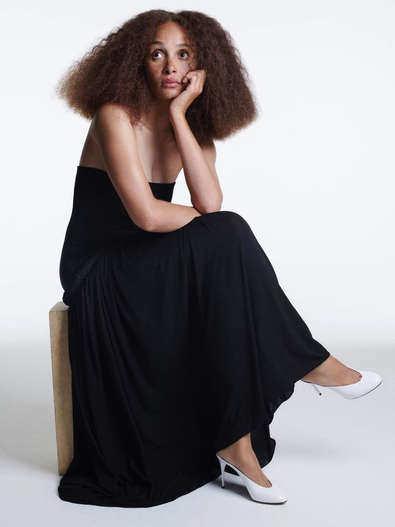 Elegant adult woman with afro sits on block wearing a black gown and white heels in a minimalist studio against a white backdrop.