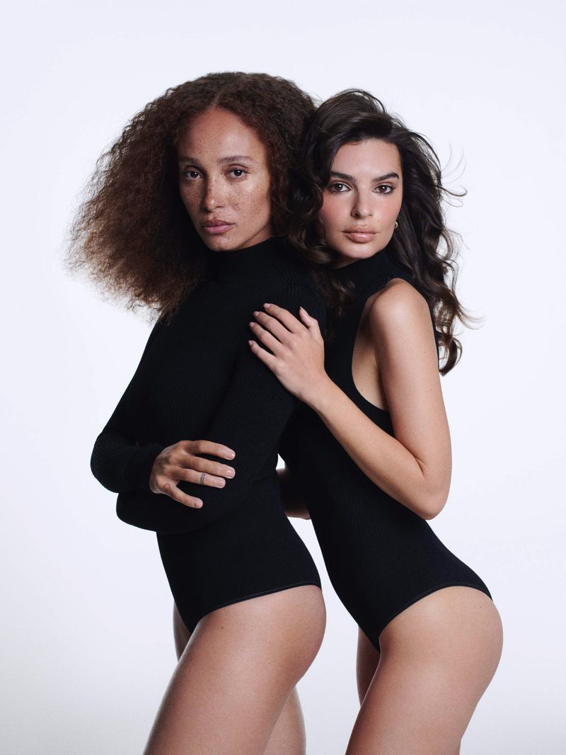 Two women in black bodysuits pose closely against a white backdrop in a studio setting, showcasing fashion and beauty.