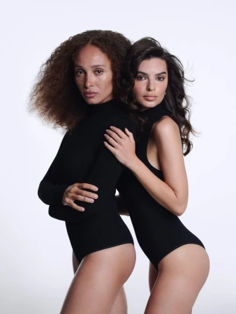 Two women in black bodysuits pose closely against a white backdrop in a studio setting, showcasing fashion and beauty.