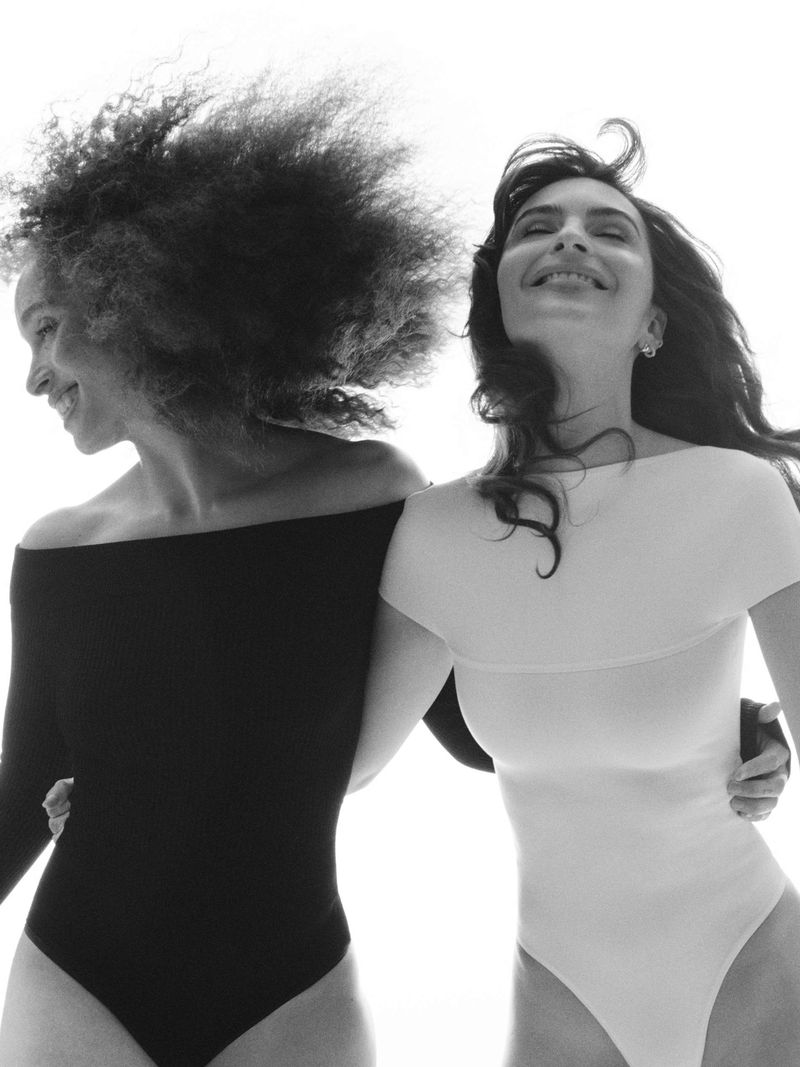 Two women in black and white bodysuits pose in a bright, high-key shot. The woman on the left has afro hair. The woman on the right has wavy hair.