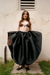 Sarah Bolger holding wide an oversized black satin balloon skirt while wearing a floral bikini top against a weathered stucco wall.
