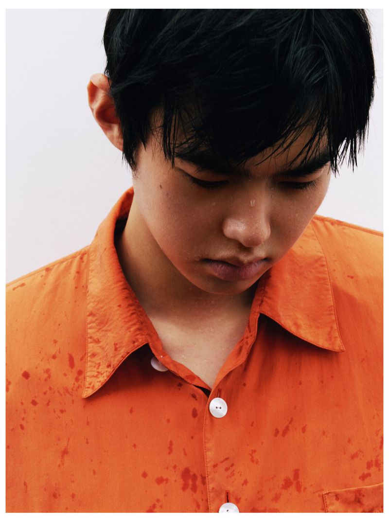 Young East Asian man looking down wearing a wet orange collared shirt against a pale overcast backdrop.
