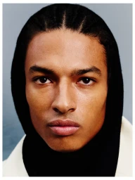 Close-up portrait of model Isak Jawo wearing a black hood and cornrows against a muted grey studio backdrop.