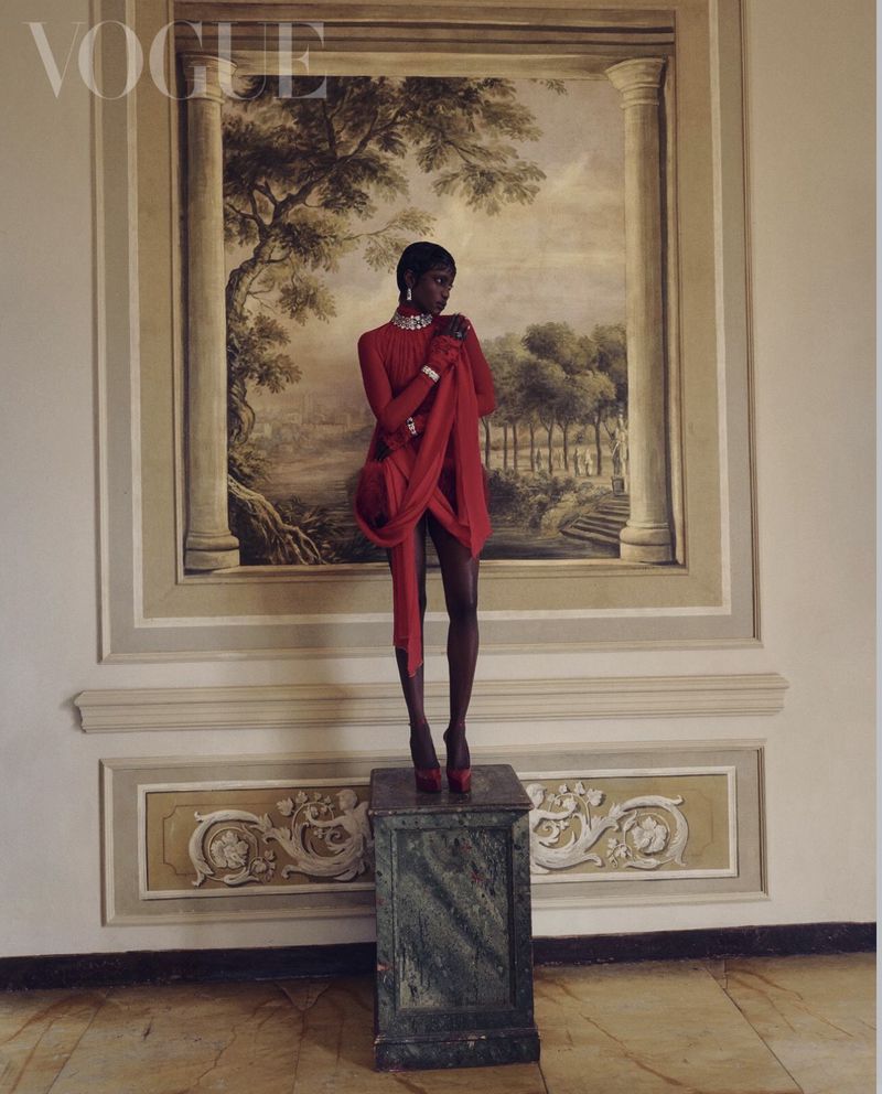 Model Rosalie Ndour wearing a red draped dress and diamond necklace stands on a pedestal before a classical landscape wall mural.