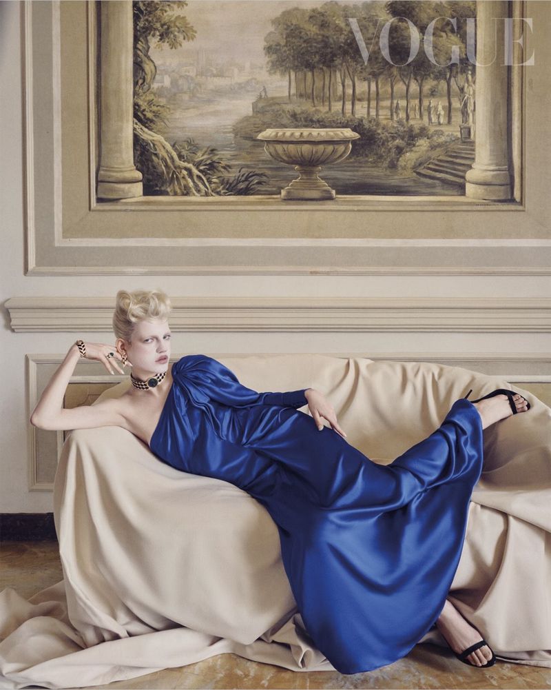 Model reclining on a draped beige sofa in a blue satin gown with Bvlgari high jewelry in front of a classical mural.