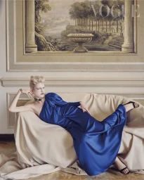 Model reclining on a draped beige sofa in a blue satin gown with Bvlgari high jewelry in front of a classical mural.