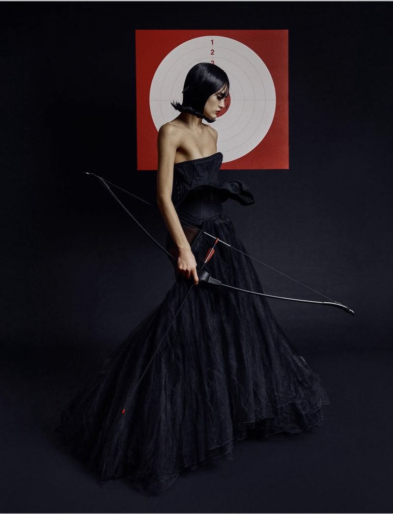 A model with a black bob hairstyle wears a strapless black evening gown, holding a bow while standing before a circular archery target.