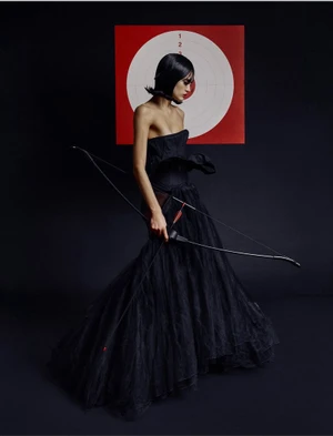 Valentina Ferrari in a black strapless tulle gown holding a bow and arrow against a black studio backdrop with an archery target.