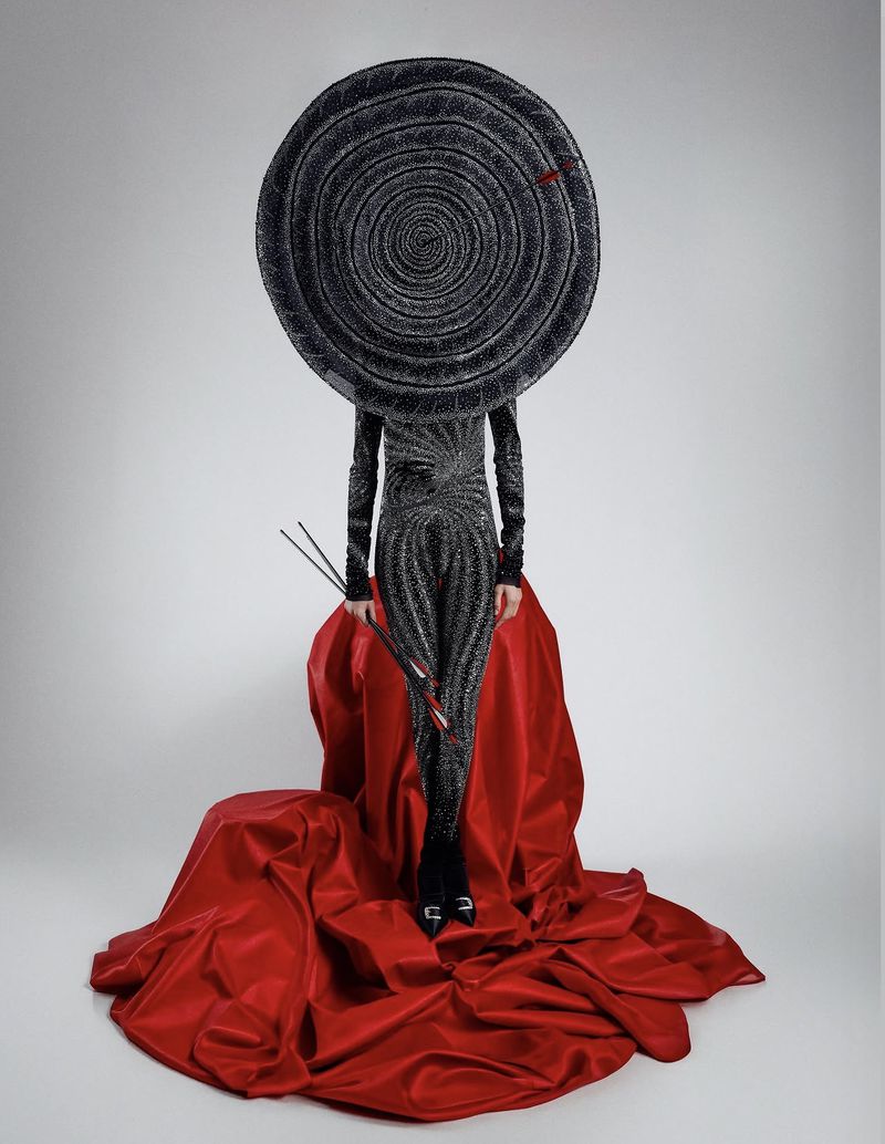A model wearing a massive circular spiral headpiece and patterned jumpsuit standing amid draped red satin fabric.