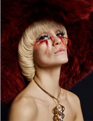 Model wearing a voluminous burgundy fur hat, blonde bowl-cut hair, red feather eyelashes, and a gold statement pendant necklace.