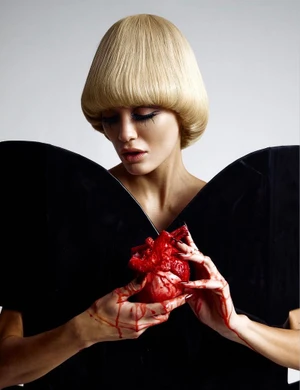 Model Valentina Ferrari with blonde bob holding a bloody anatomical heart wearing a black sculptural top.