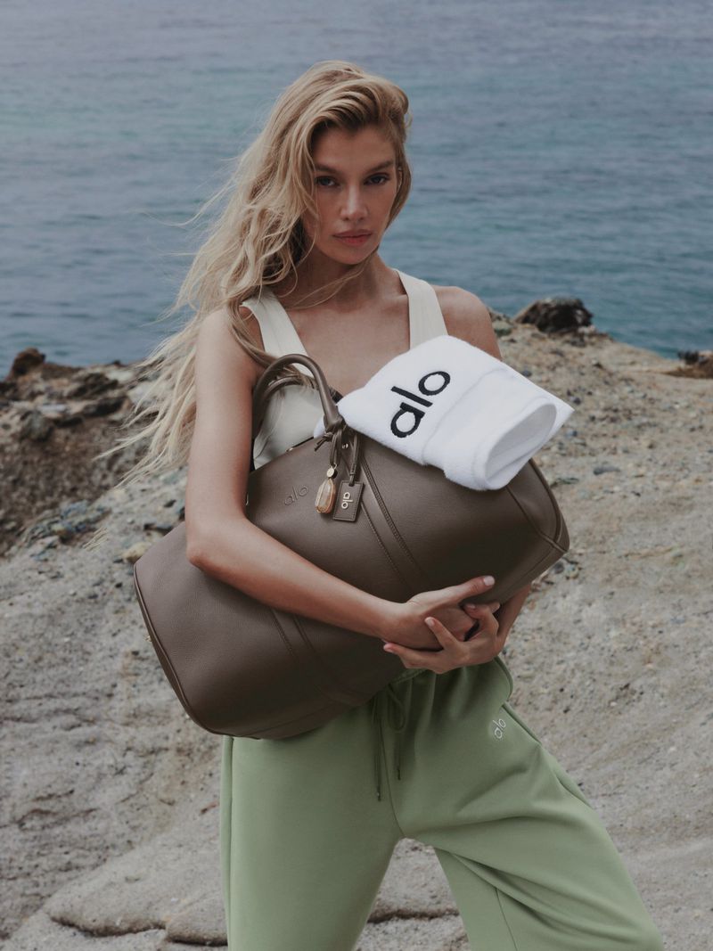 A blonde woman in a white tank top and sage green joggers holds a large brown leather duffel bag and a white towel with Alo logo.