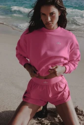 Daiane Sodre kneeling on a sandy beach wearing a bright pink Alo sweatshirt and matching athletic shorts.