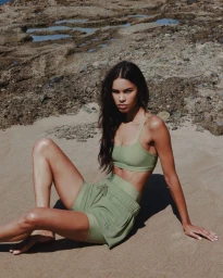 Model Daiane Sodre seated on a sandy beach wearing a matching sage green sports bra and athletic shorts set for Alo.
